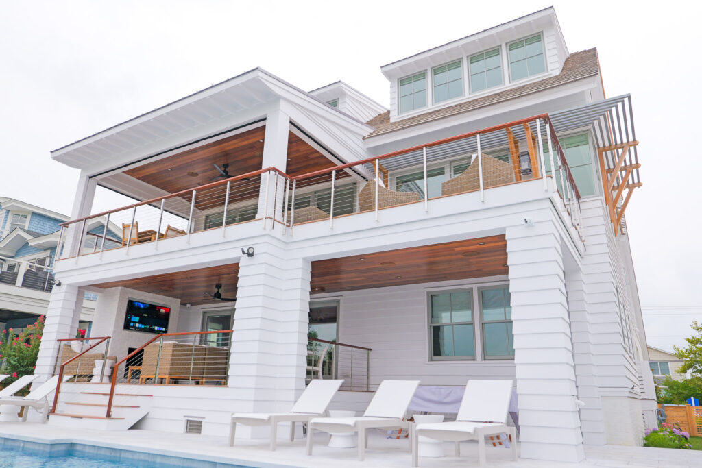 The rear of a large house with a pool, outdoor deck on the first and second floor, and an outdoor TV and entertainment area. Custom Home Technology Integration includes outdoor entertainment