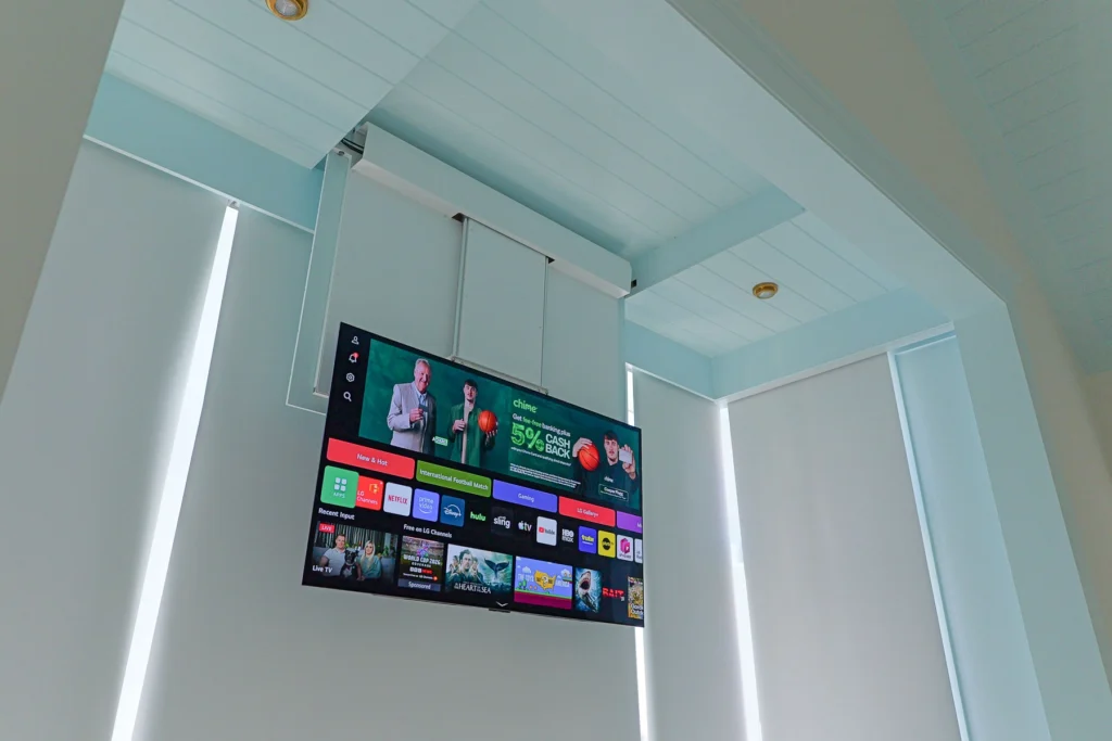 Ceiling-mounted drop-down TV deployed from the alcove ceiling in a Stone Harbor, NJ primary bedroom, with Lutron motorized shades visible behind, installed by Cooltronics.